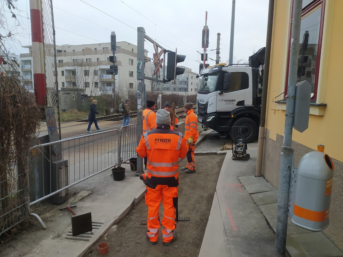 Pfnier-Bauarbeiter führen Arbeiten an der Eisenbahnkreuzung durch, während ein LKW der Firma bereitsteht. Der laufende Zugverkehr wird dabei nicht beeinträchtigt.