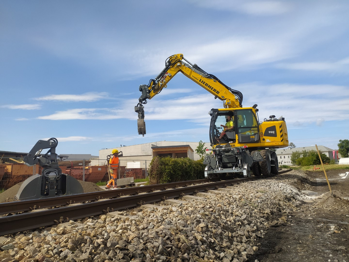 Ein moderner Schienenbagger von Liebherr hebt eine schwere Betonplatte auf einer Baustelle an, während ein Arbeiter in orangefarbener Schutzkleidung die Arbeiten überwacht.