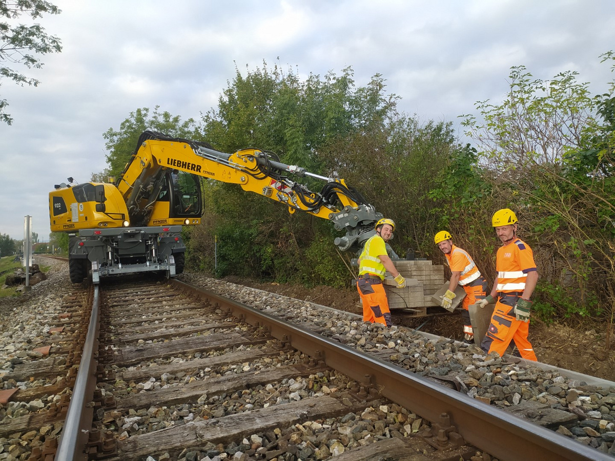 Drei Arbeiter in orangefarbener Schutzkleidung arbeiten zusammen mit einem Schienenbagger an der Verlegung von Kabelkanälen entlang der Bahngleise.