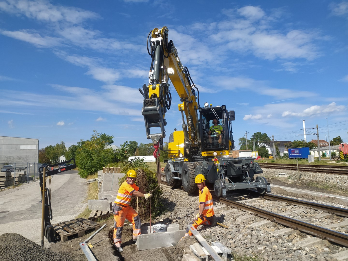 Ein Liebherr Schienenbagger hebt eine Betonplatte, während zwei Arbeiter in Schutzkleidung Vorbereitungen entlang der Schiene treffen.