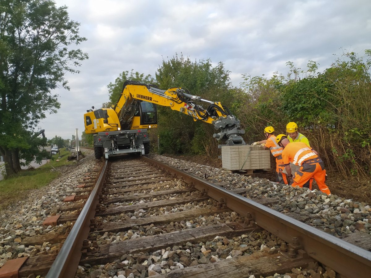 Ein Schienenbagger unterstützt drei Pfnier-Arbeiter beim Entladen schwerer Betonplatten entlang der Gleise.