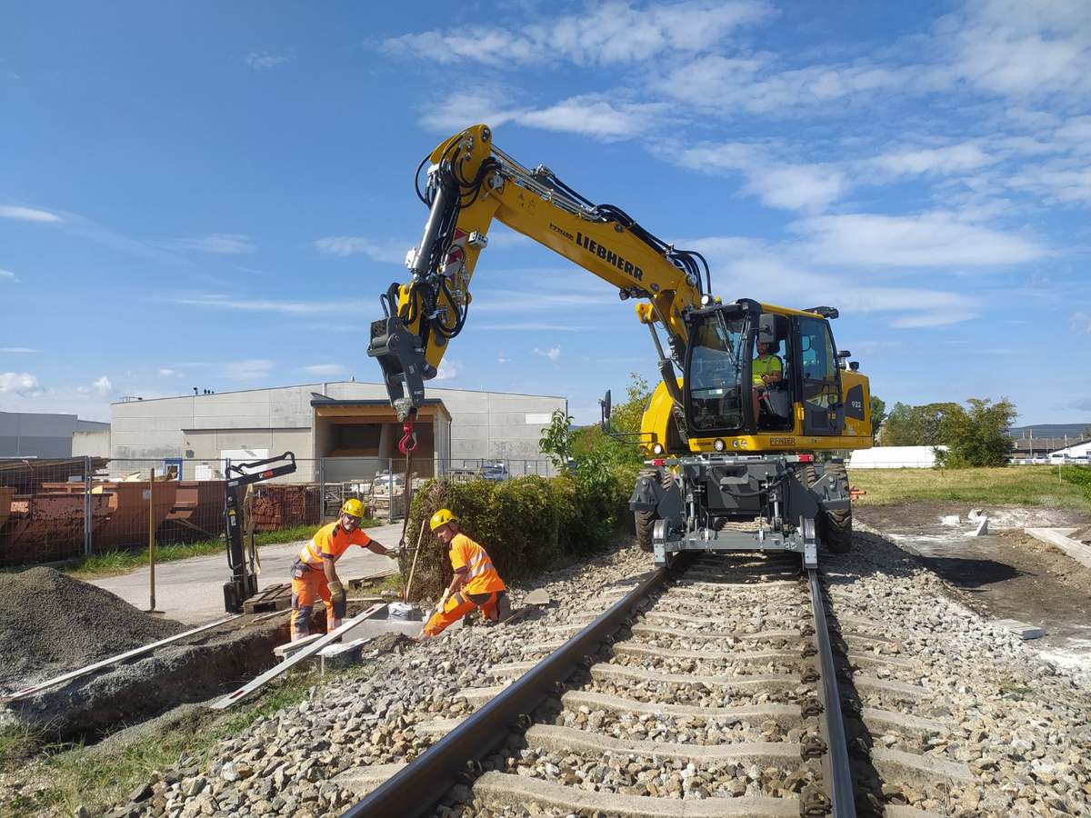 Zwei Arbeiter in Sicherheitskleidung arbeiten an einer Trogtrasse entlang einer Schienenstrecke, unterstützt von einem Liebherr Schienenbagger, der eine Last hebt.